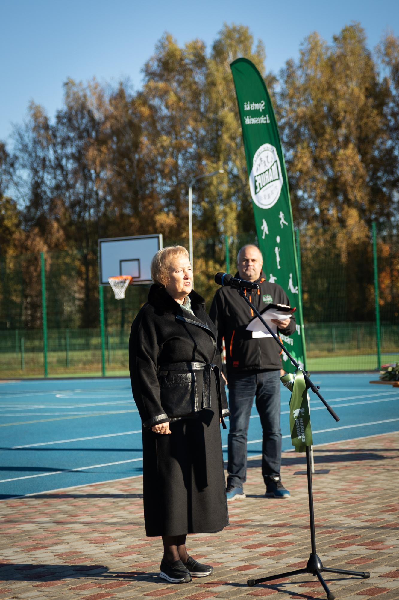 Sieviete tumšās drēbēs stāv pie mikrofona statīva sporta laukumā