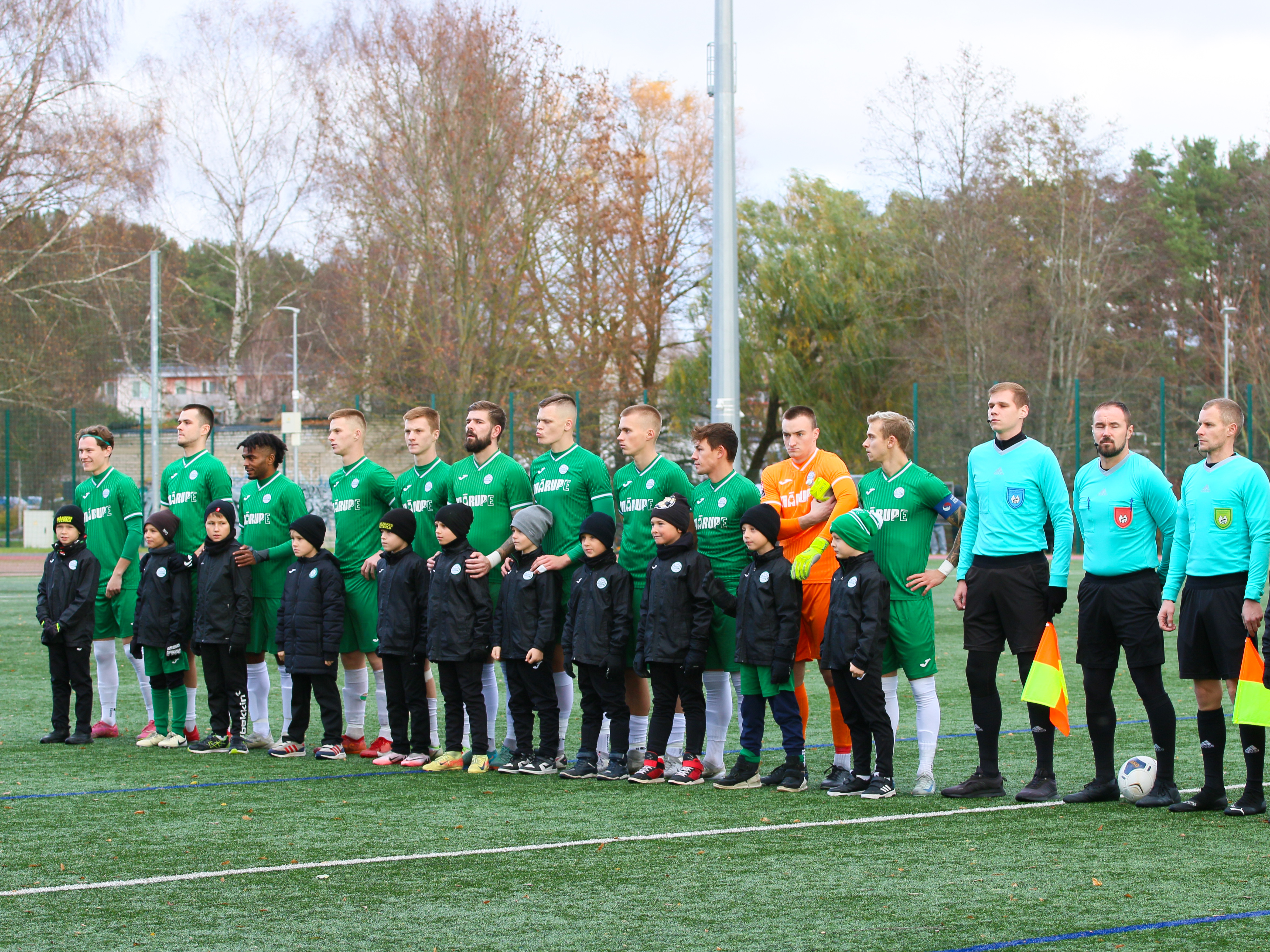 Futbolisti un spēles tiesneši stāv vienā rindā futbola laukumā