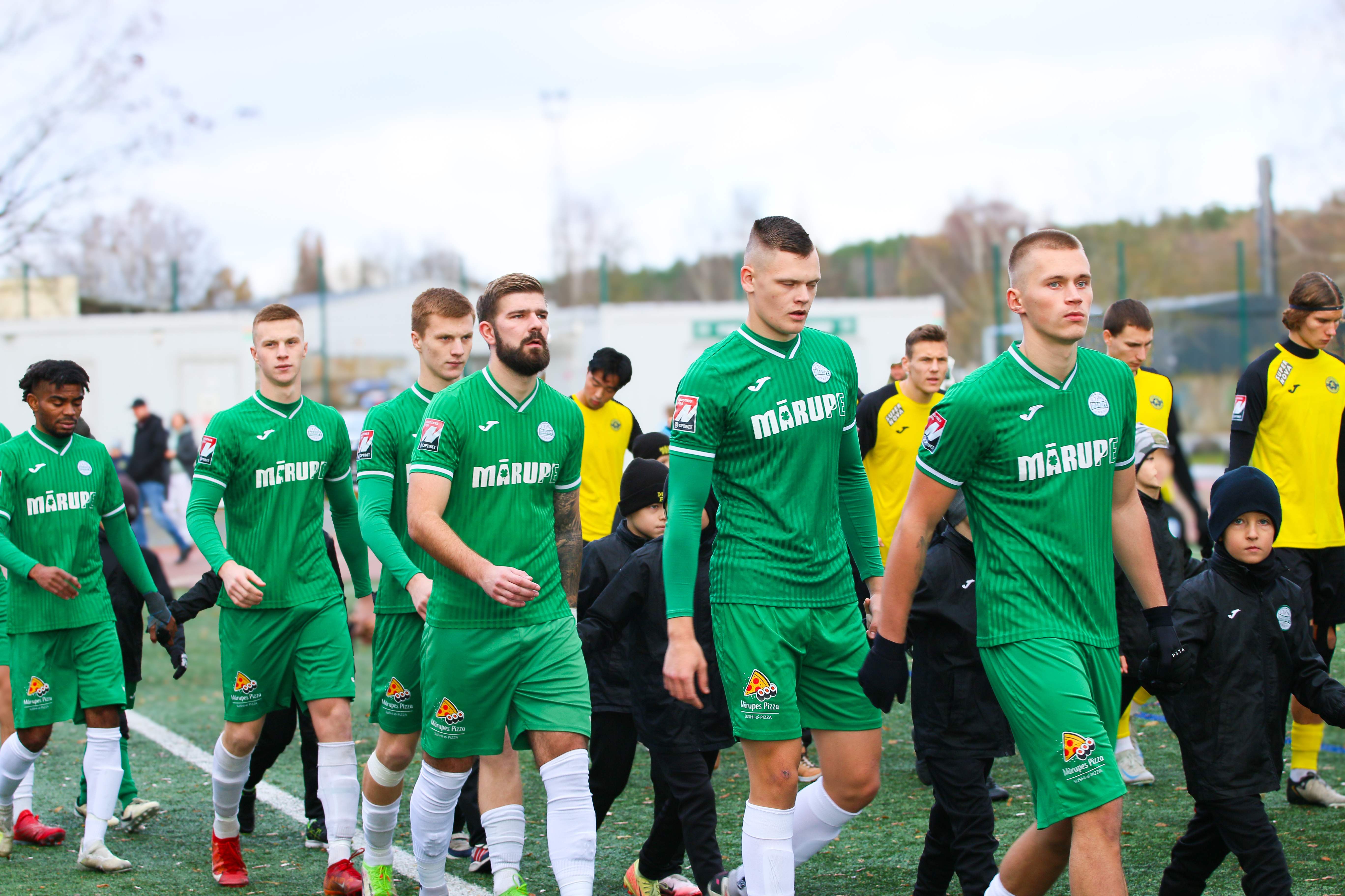 Vairāki futbola spēlētāji zaļās sporta drēbēs iznaķ futbola laukumā 