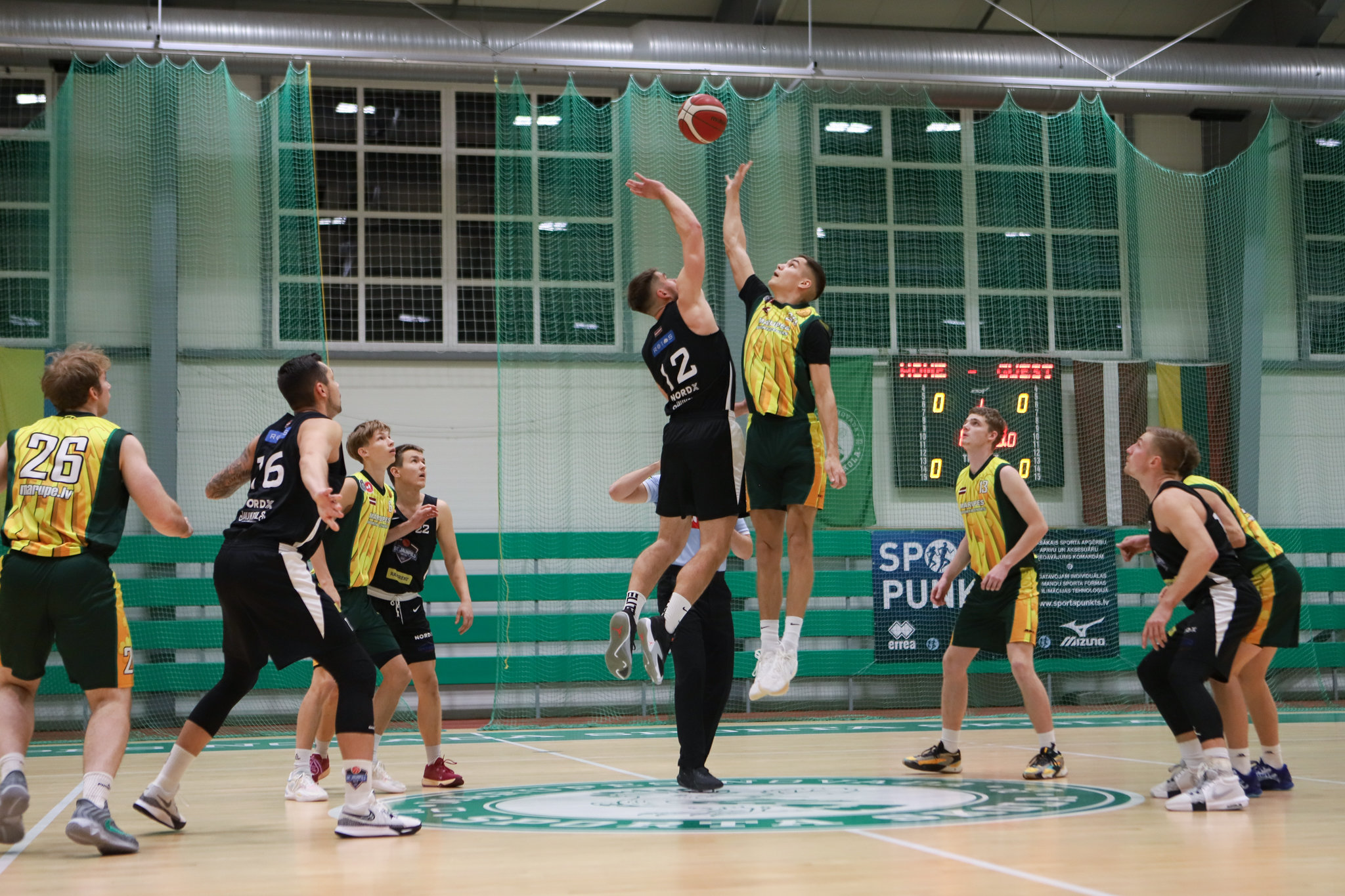 Basketbolisti melnos un dzeltenos tērpos cīnās par basketbola bumbu sporta zālē