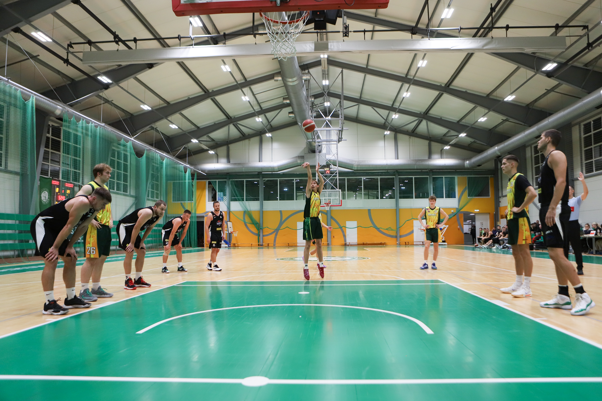 Basketbolists dzeltenā kreklā stāv pretī grozam un bet bumbu, sānos basketbolisti melnos tērpos