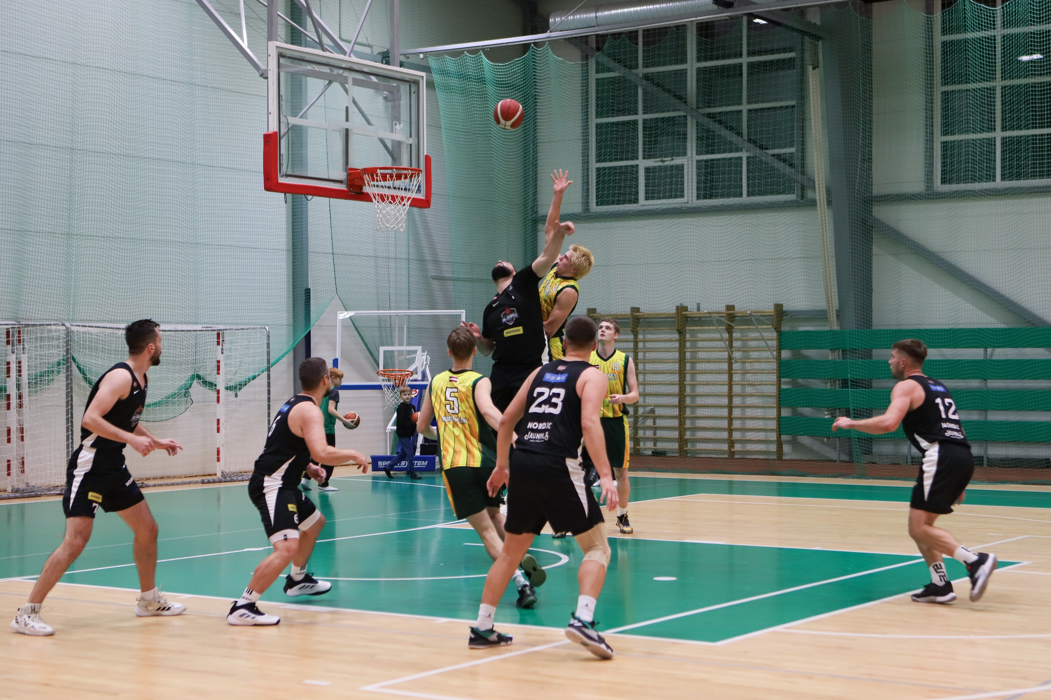 Basketbolisti melnos un dzeltenos tērpos cīnās par basketbola bumbu sporta zālē