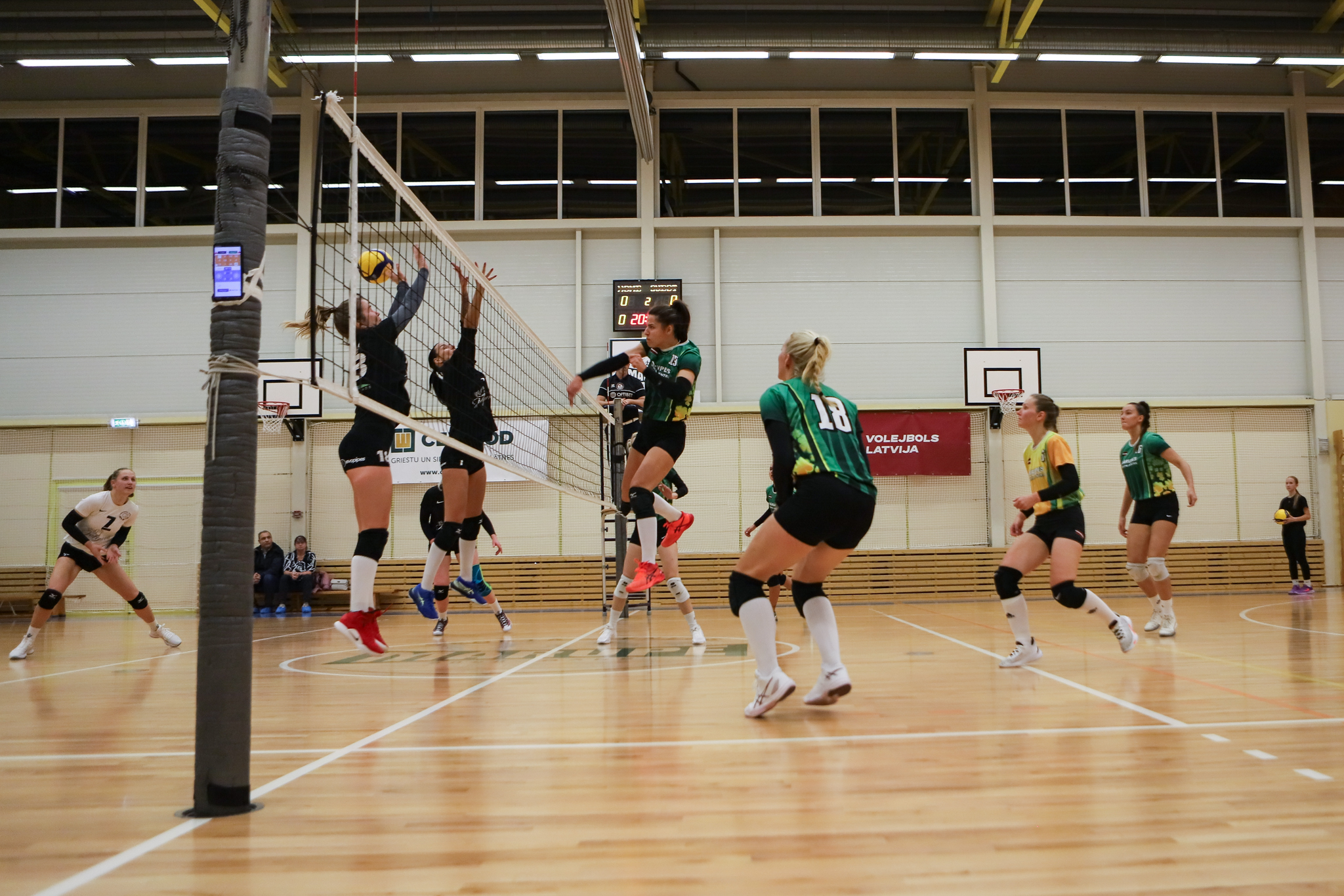 Sieviete zaļā sporta tērpā sit pāri volejbola bumbu pāri tīklam, kur to otrā laukuma pusē cenšas notvert sievietes melnos sporta tērpos