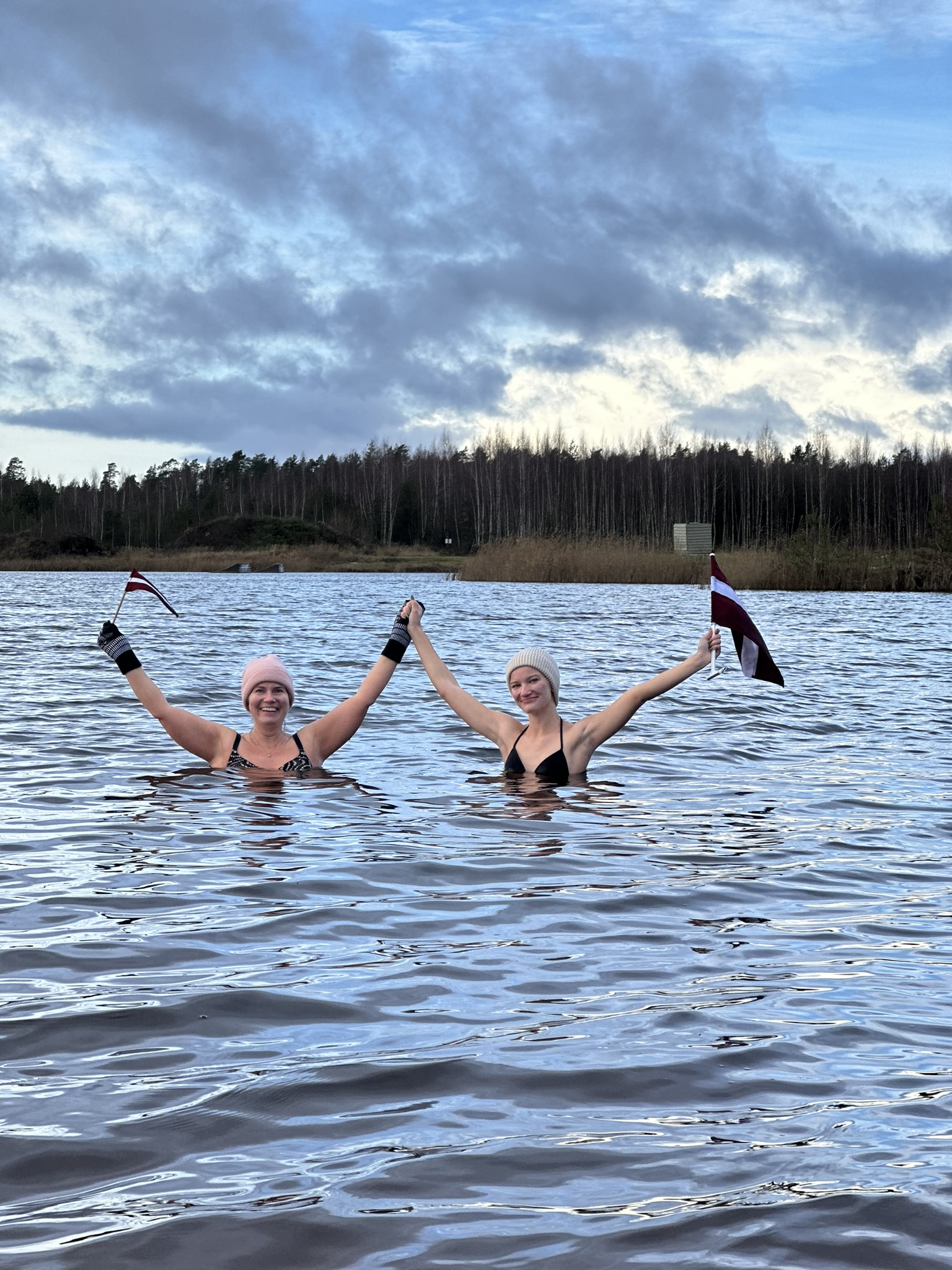 Divi cilvēki ūdenstilpnē stāv un tur Latvijas karogi