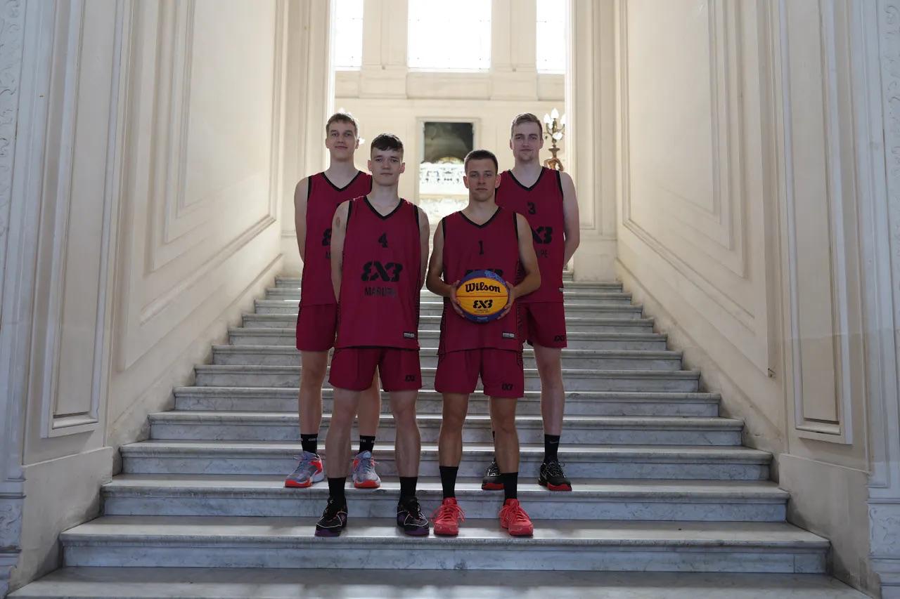 Četri puiši sarkanos sporta tērpos stāv uz baltām trepēm, vienam no puišiem rokās basketbola bumba