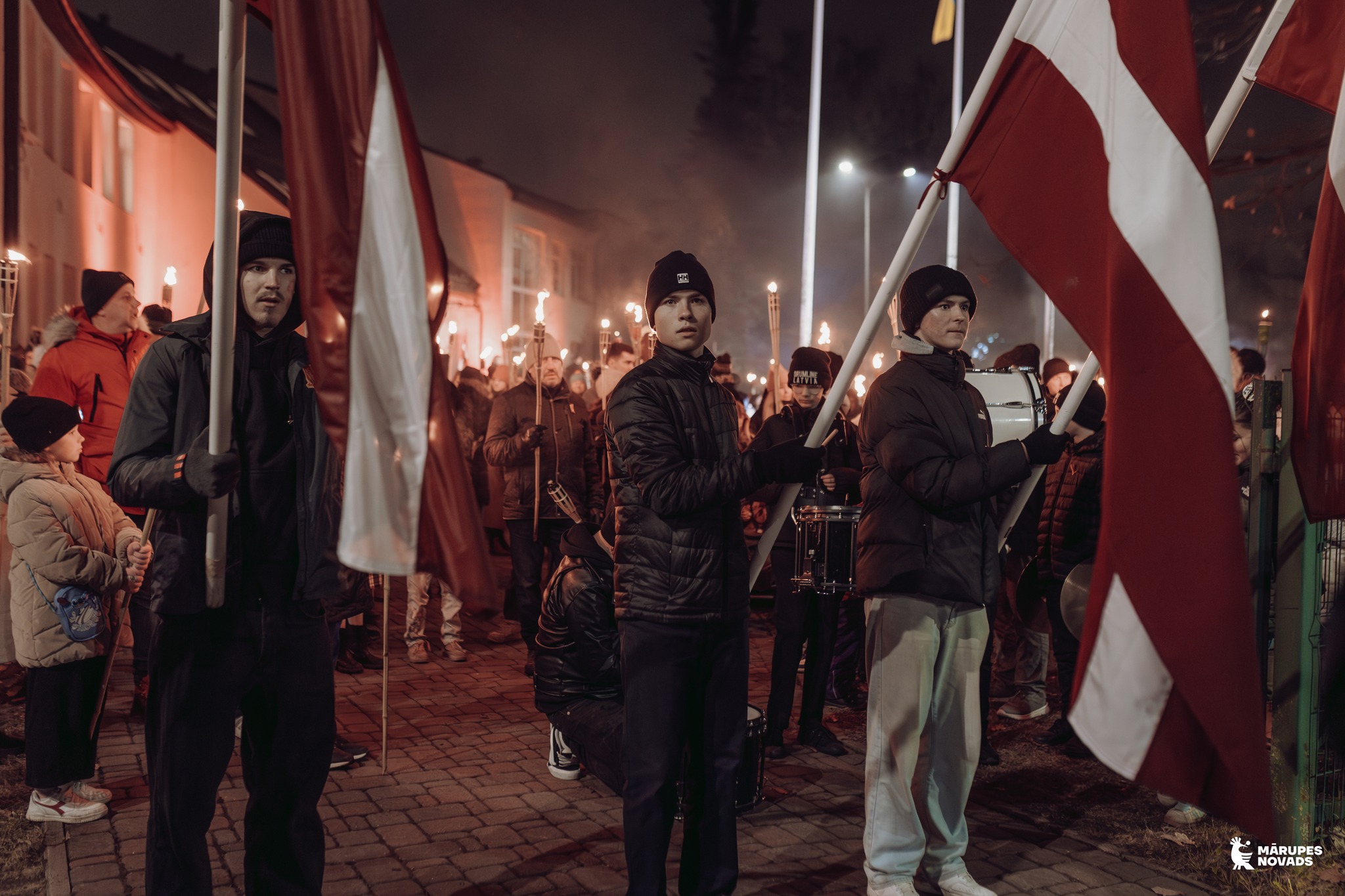 Jaunieši stāv tumsā ar Latvijas karogiem rokās pirms Lāpu gājiena