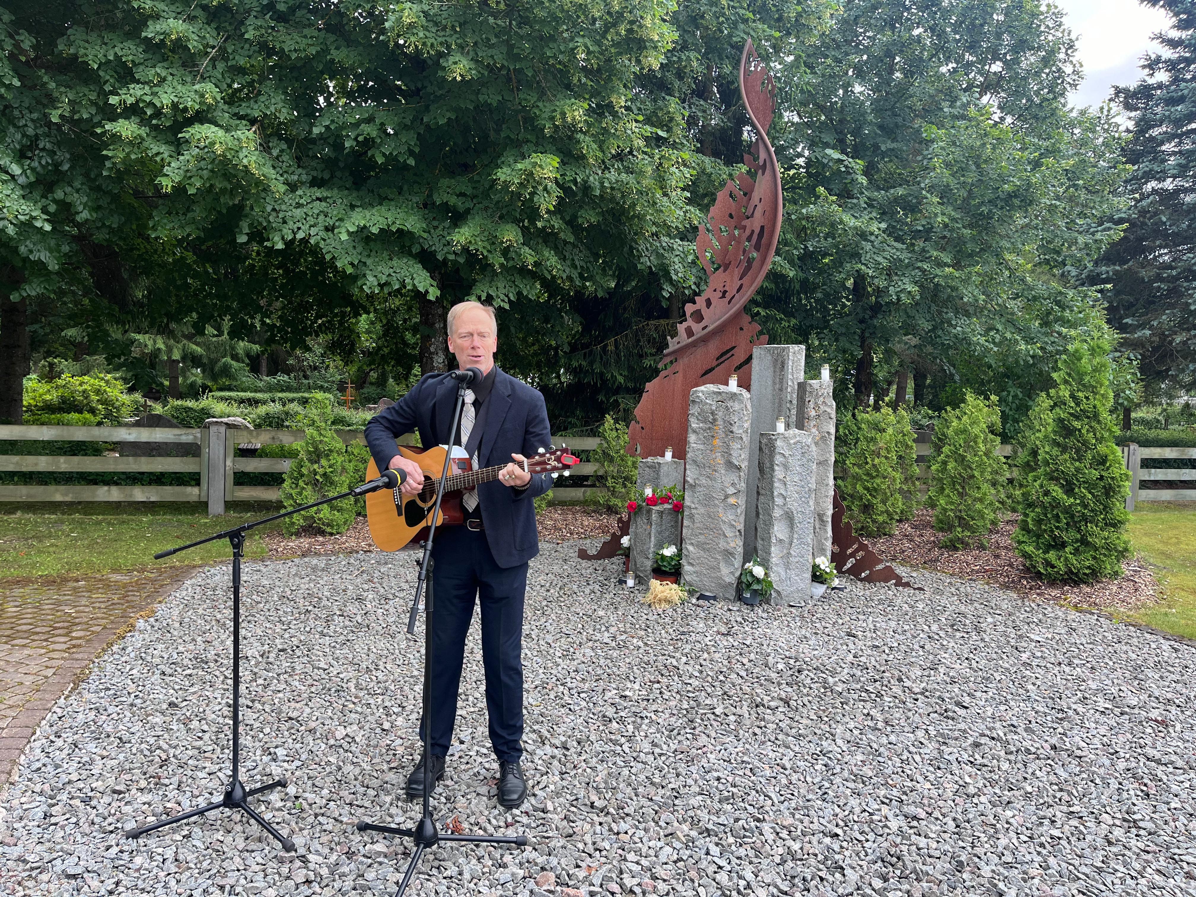 Vīrietis ar ģitāru rokās stāv pie pieminekļa un dzied