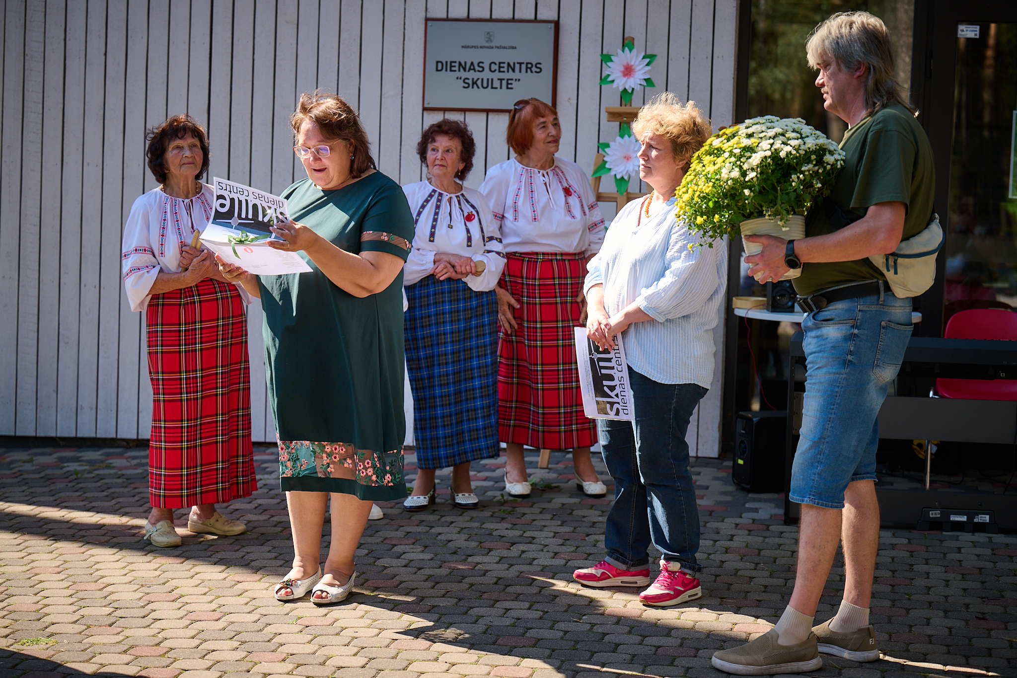 Cilvēki pie ēkas ar ziediem rokās saka runu pasākumā