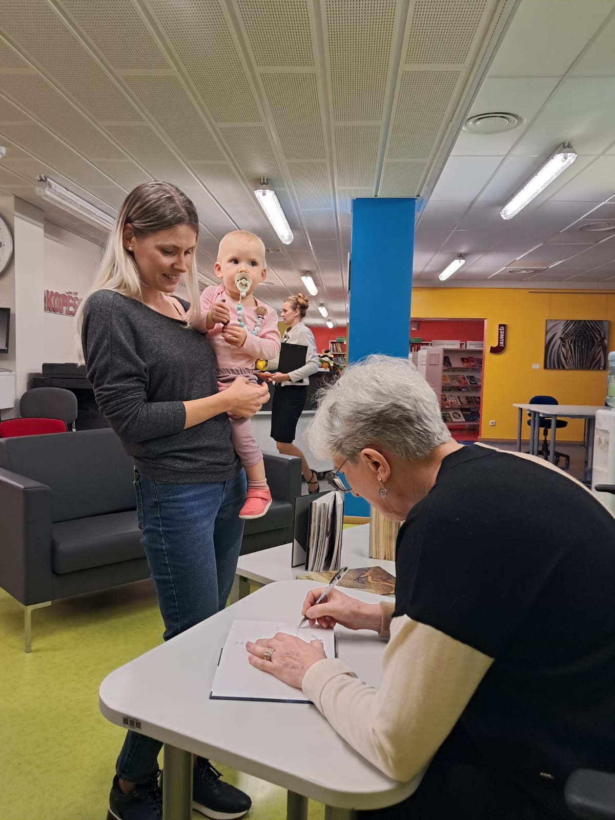 Sieviete ar mazu bērniņu rokās stāv rindā, lai saņemtu grāmatas autores autogrāfu