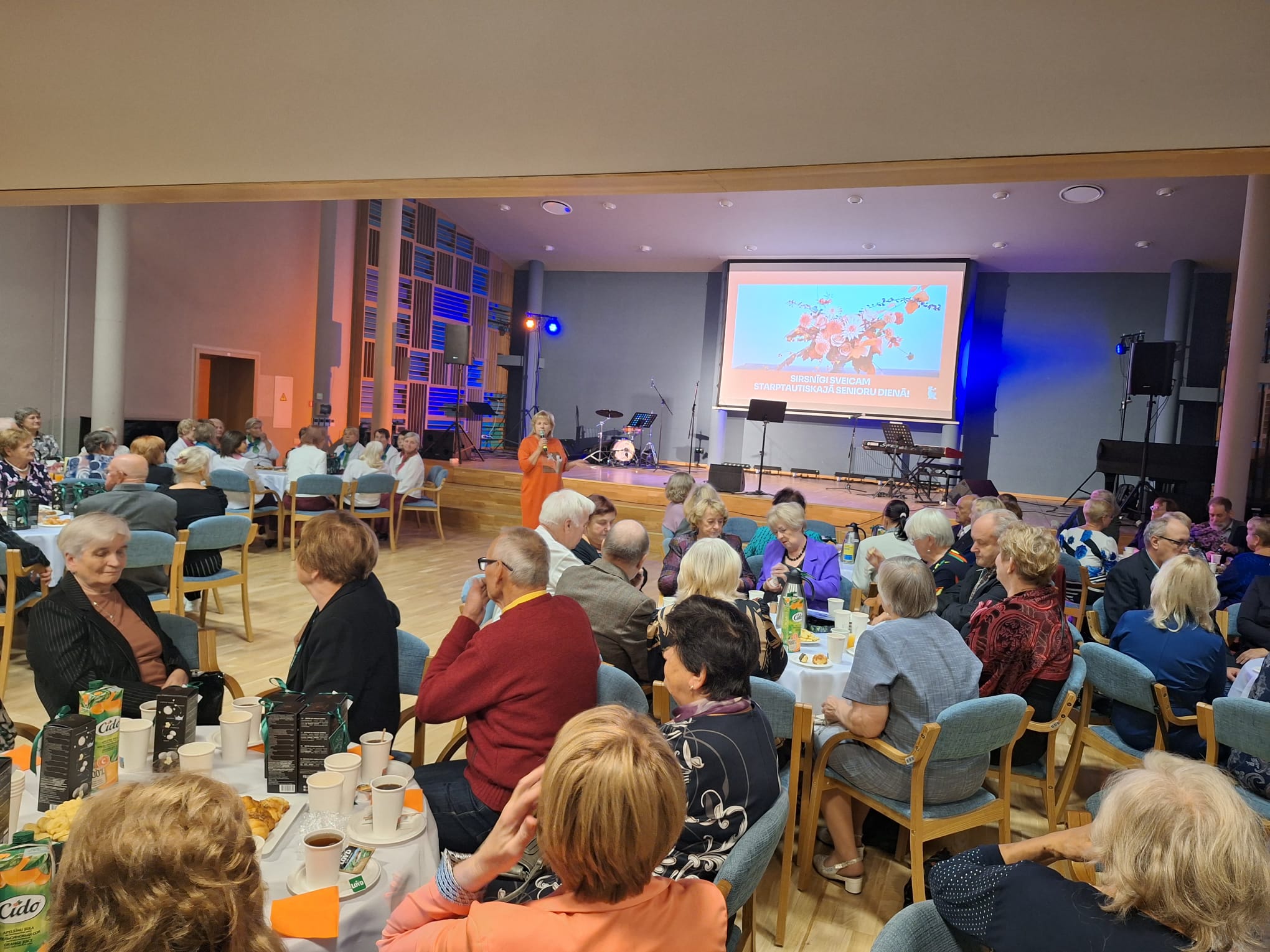 Pasākumu zāle ar dalībniekiem pie galdiņiem