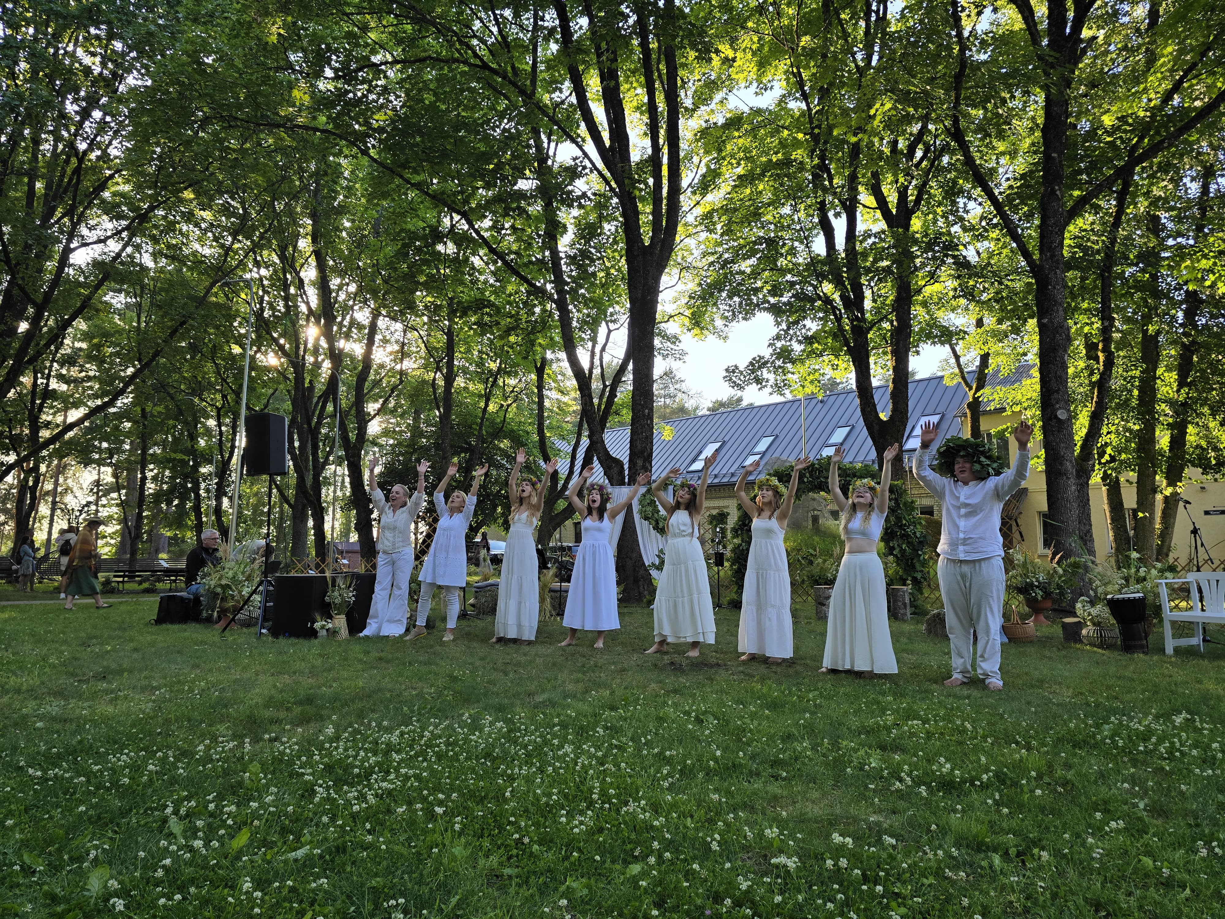 Popgrupas dalībnieki baltos kostīmos dzied un dejo saulainā vasaras vakarā brīvdabā
