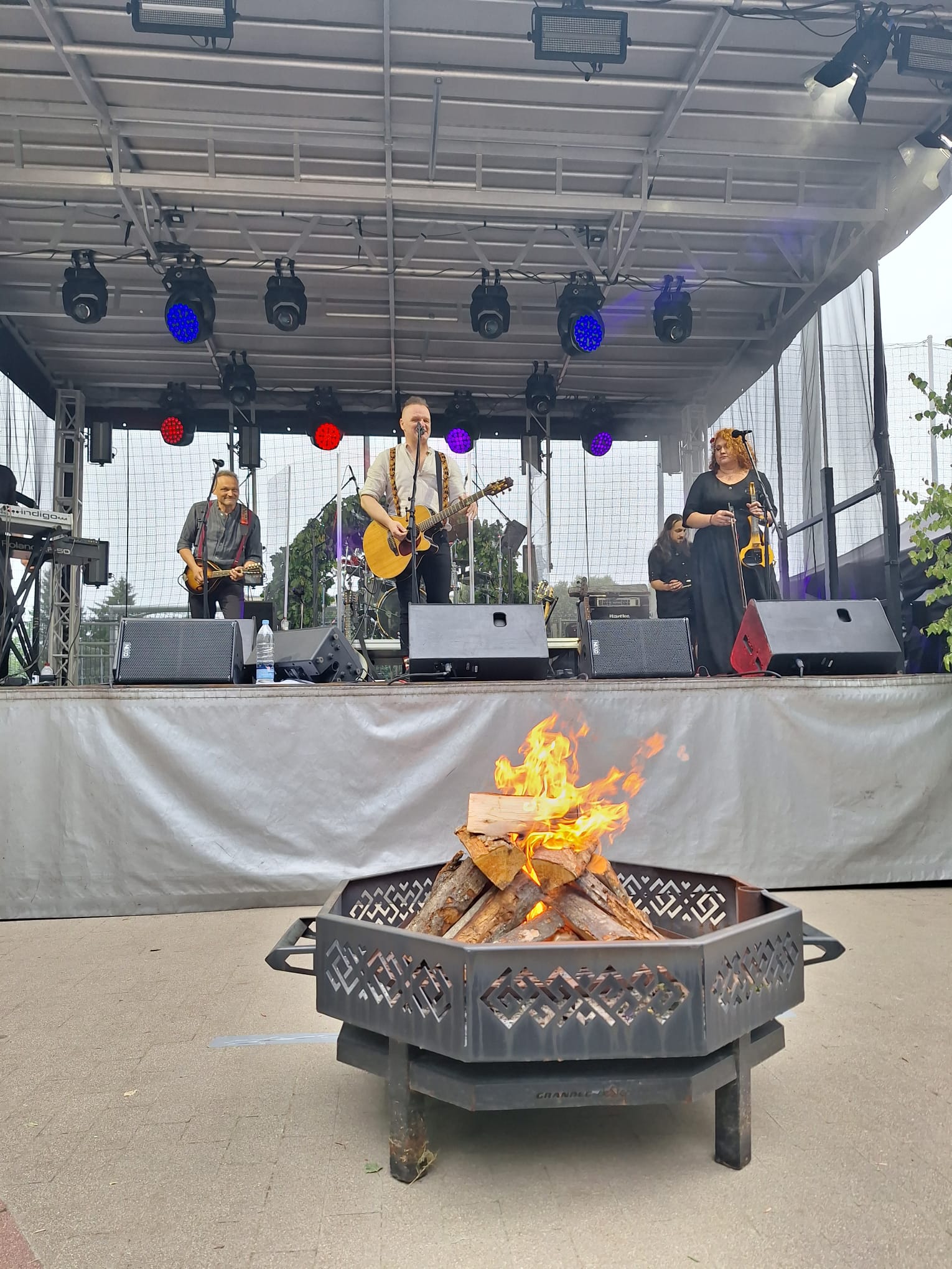 Mūzikas grupas dalībnieki uz skatuves, priekšplānā deg ugunskurs