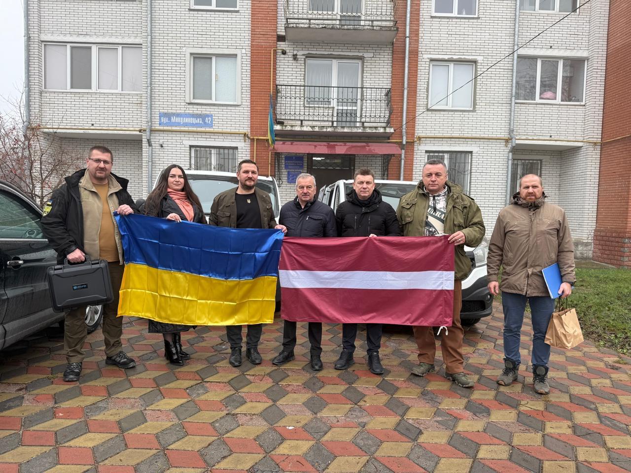 Cilvēku grupa pie dzīvokļu nama ar Latvijas un Ukrainas karogiem rokās