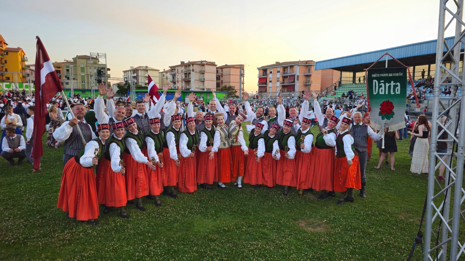 Deju kolektīva dalībnieki balti sarkanos tērpos ar karogiem stadionā pēc festivāla gājien