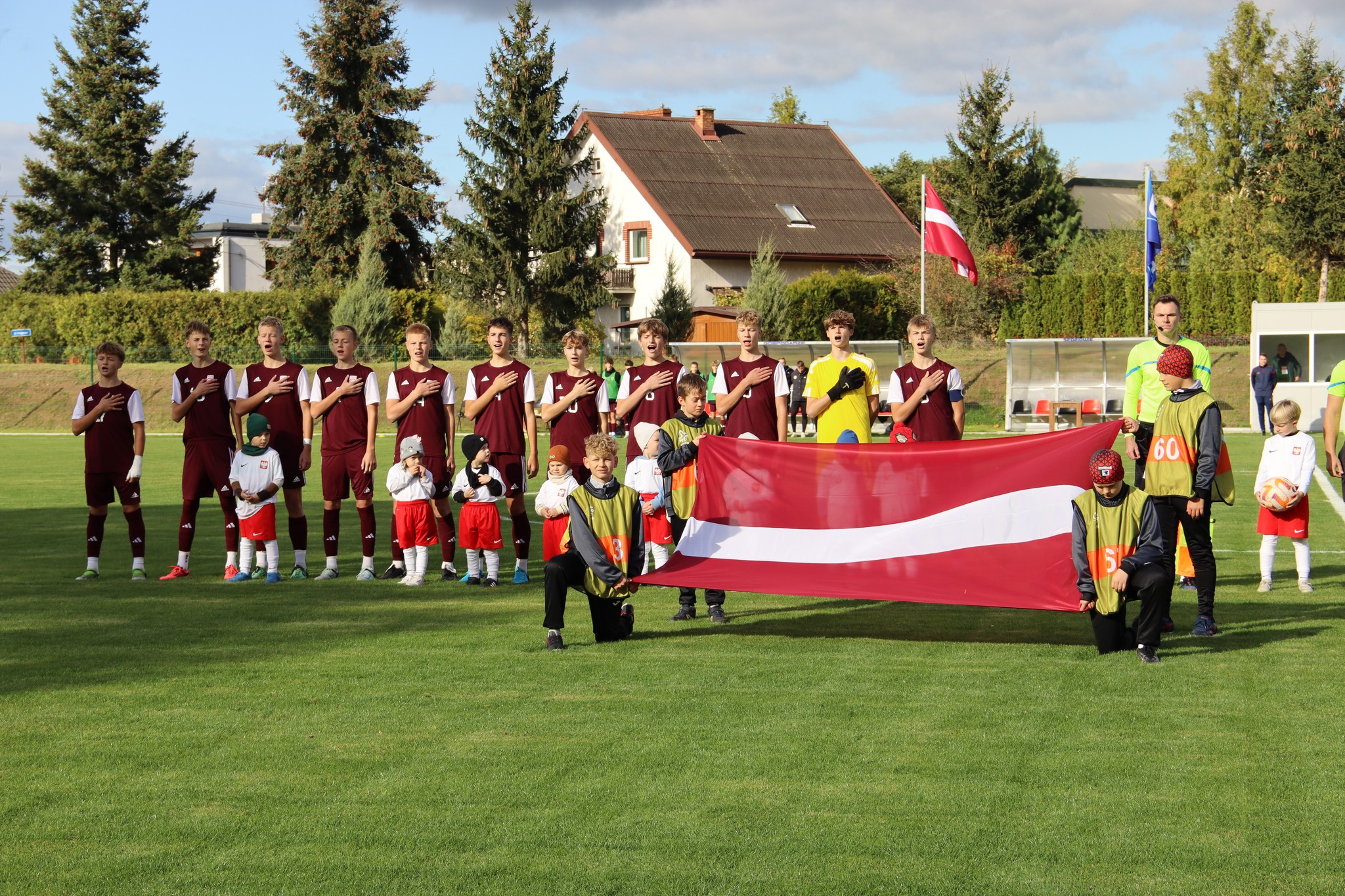 Divas komandas ar futbolistiem stāv futbola laukumā kur priekšā bērni tur Latvijas karogu