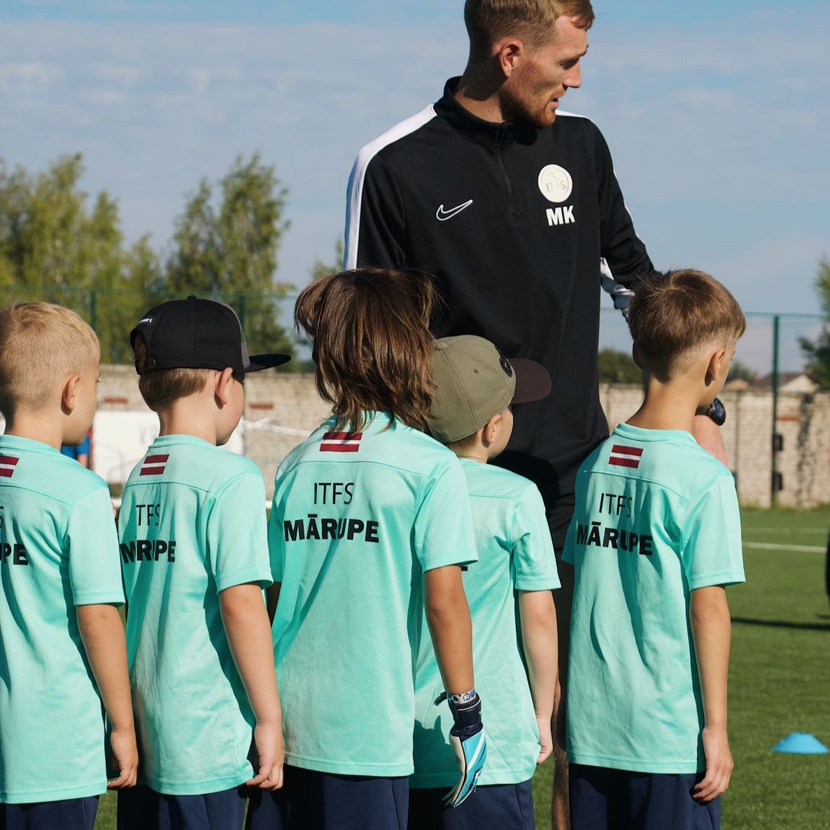 Mazi bērni zilos kreklos kopā ar vīrieti melnā jakā futbola laukumā