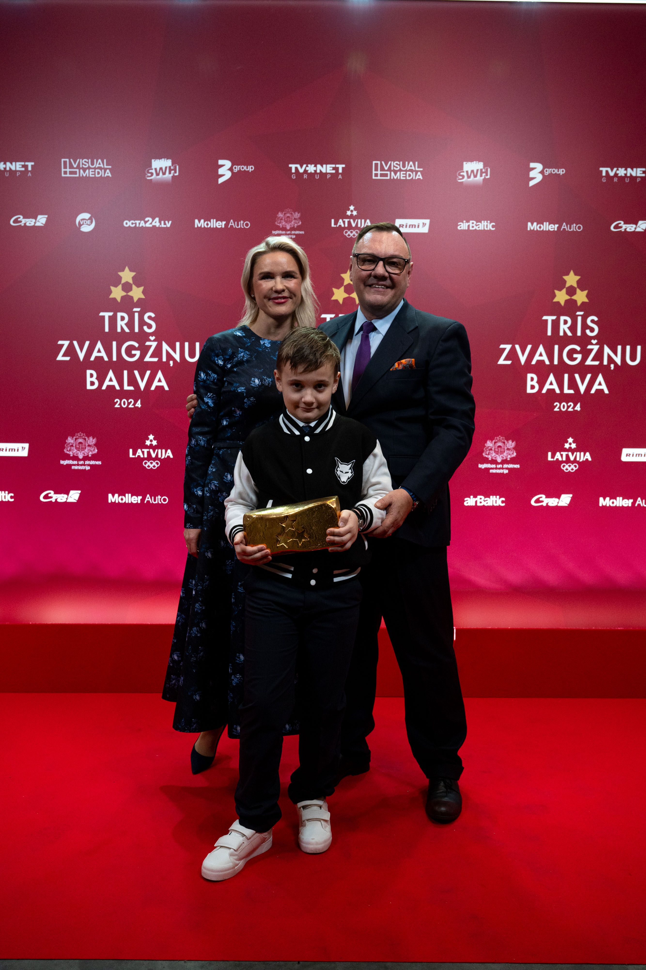 Sieviete zilā kleitā  un blondiem matiem kopā ar vīrieti uzvalkā un bērnu, kurš tur rokās zelta ķiegeļi, trofeju.