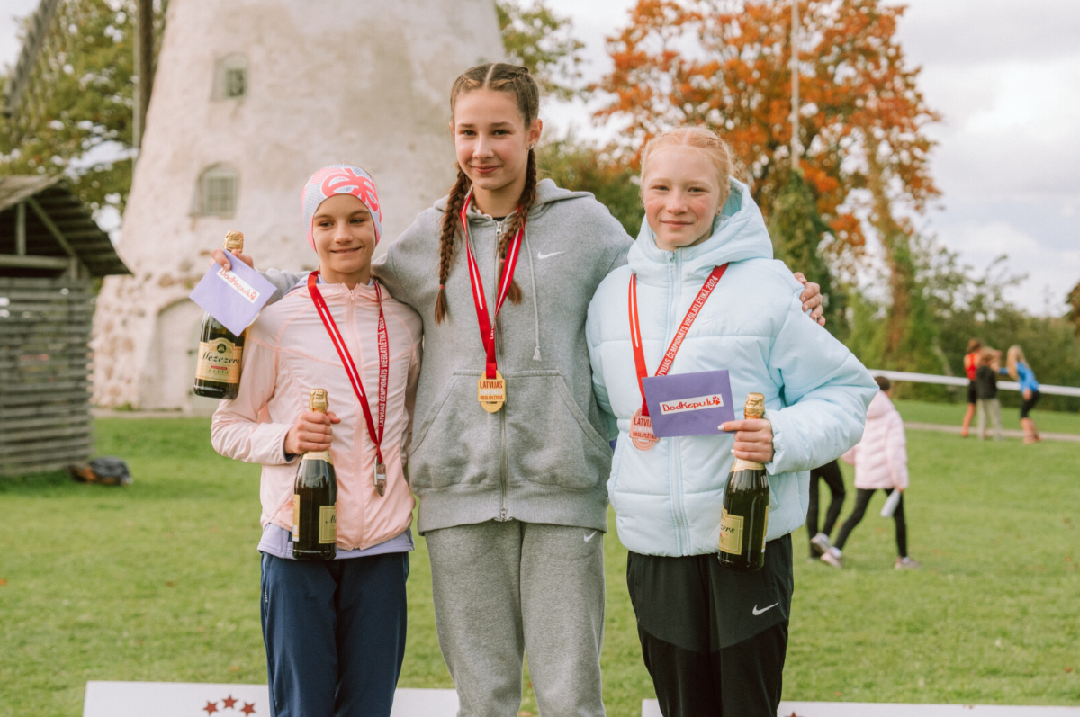 Trīs meitenes stāv uz pjedestāla ar medaļām kaklā