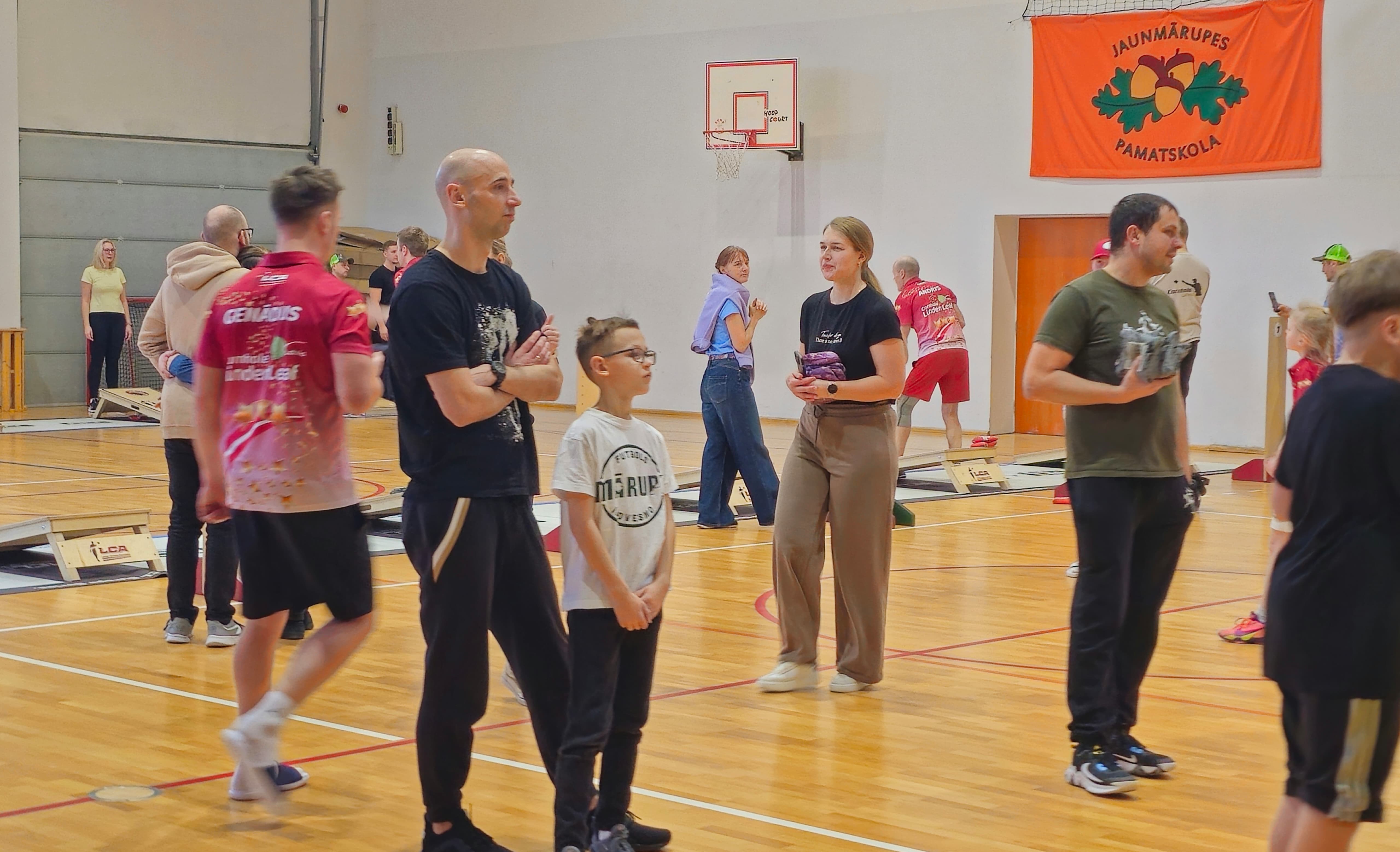 Cilvēki sporta zālē met auduma maisiņu uz koka plātnes