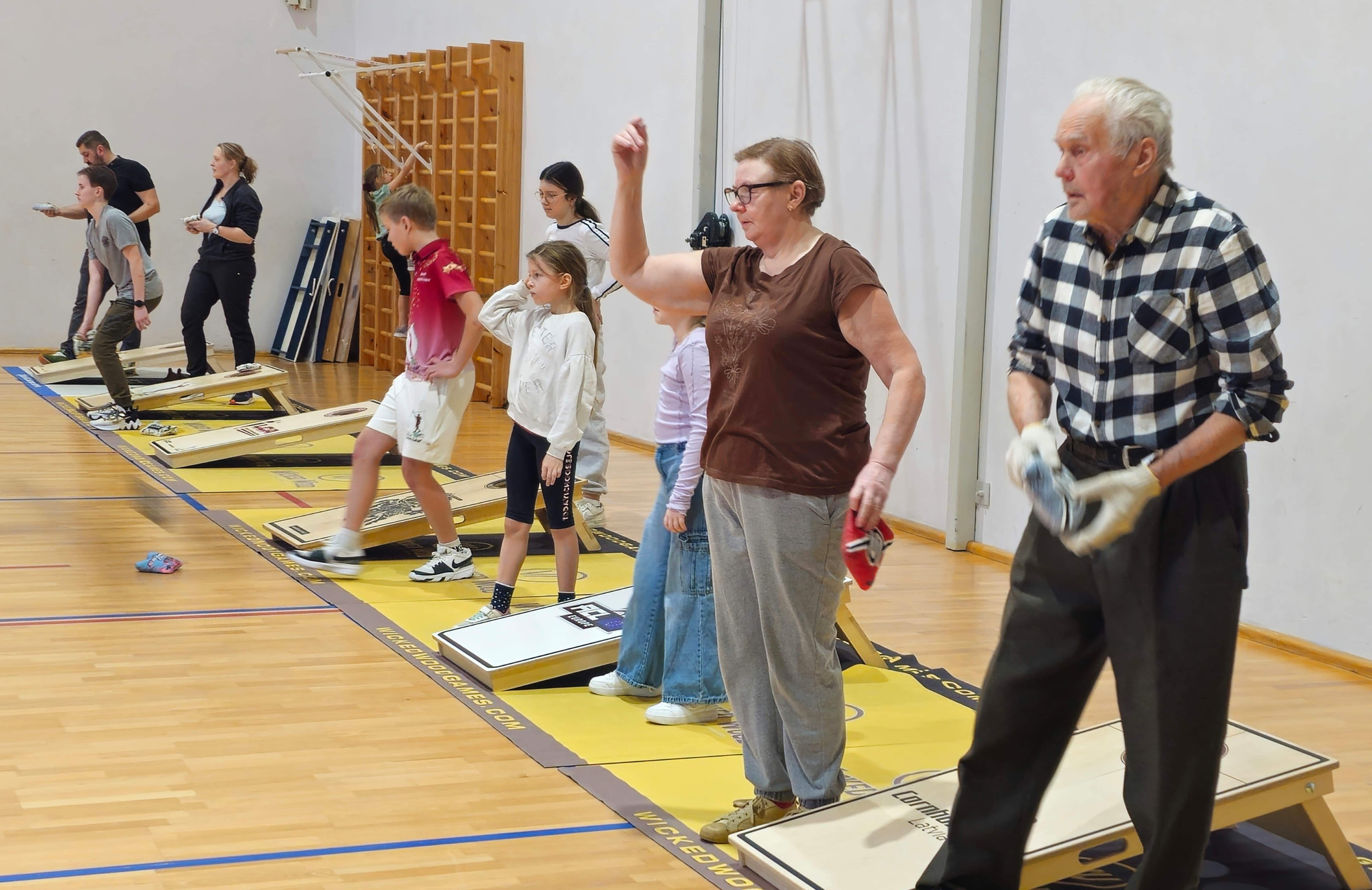 Cilvēki sporta zālē met auduma maisiņu uz koka plātnes