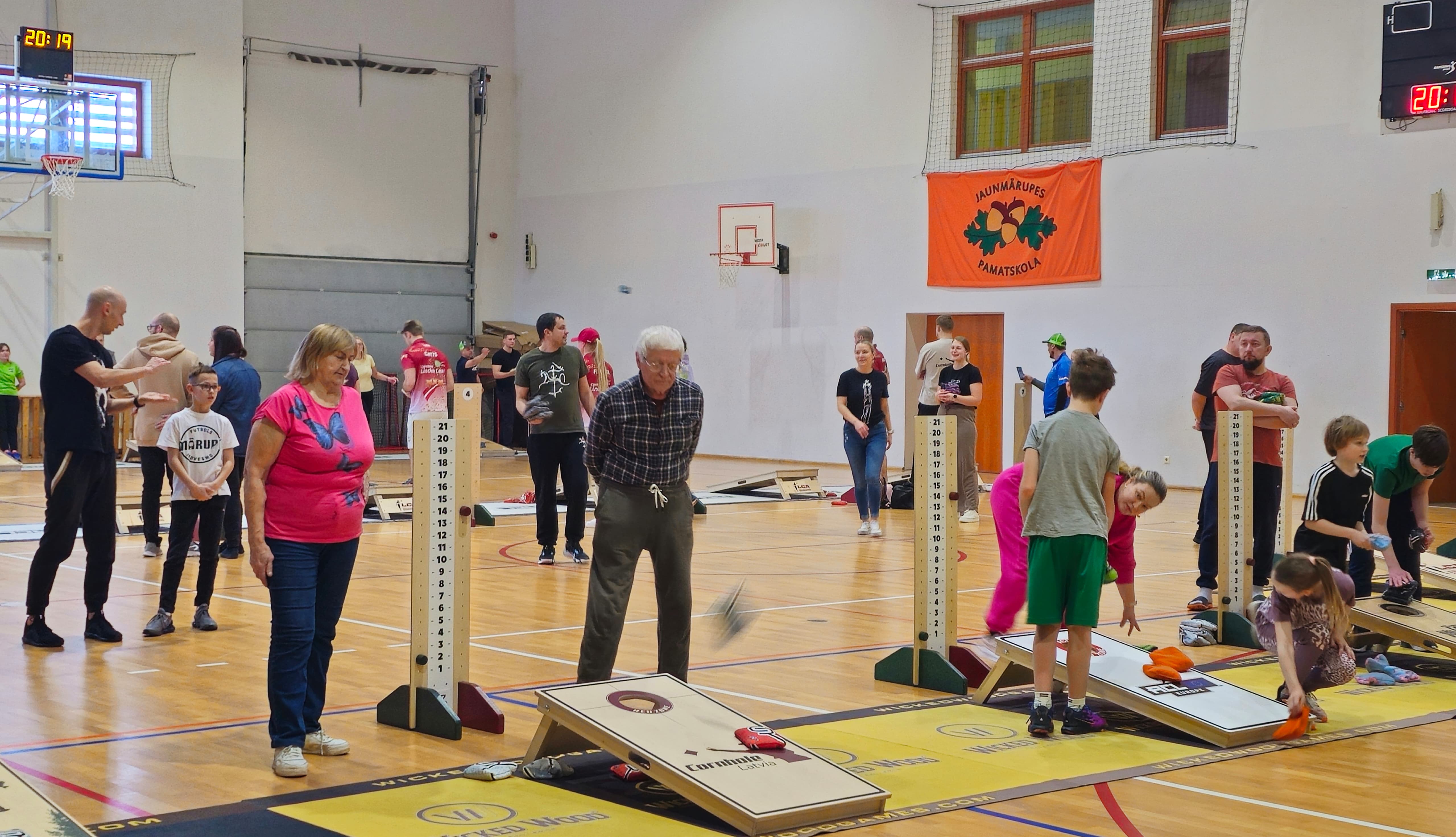 Cilvēki sporta zālē met auduma maisiņu uz koka plātnes