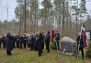 Bērnu koris tumšās drēbēs stāv mežā pie piemiņas akmens ar Latvijas karogiem un dzied