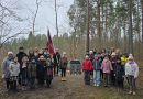 Bērnu kora dalībnieku kopbilde mežā pie piemiņas akmens ar Latvijas karogiem