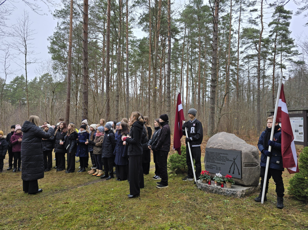 Bērnu koris tumšās drēbēs stāv mežā pie piemiņas akmens ar Latvijas karogiem un dzied