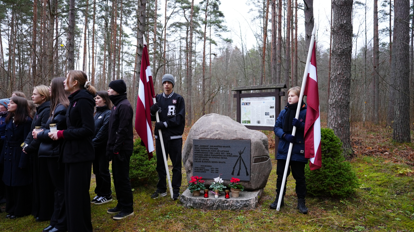 Cilvēku pulks tumšās drēbēs stāv mežā pie pieminekļa ar Latvijas karogiem