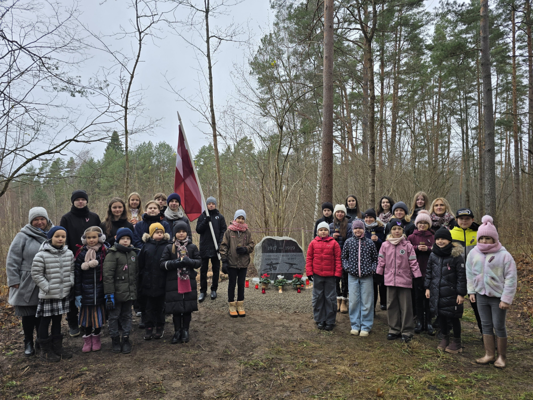 Bērnu kora dalībnieku kopbilde mežā pie piemiņas akmens ar Latvijas karogiem