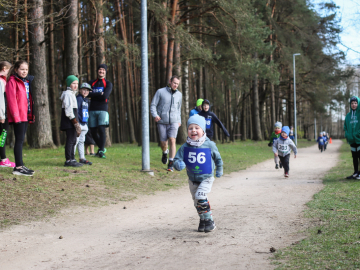 Bērns sporta tērpā skrien pa ceļu, kur gar malu stāv pieaugušie