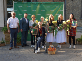 Parasportists Rihards Snikus kopā ar komandu un domes priekšsēdētāju Andreju Enci un priekšsēdētāja vietnieku Valdi Kārkliņu ar ziediem un dāvamām rokās atrodas pie zaļa foto banera, uz kura ir Mārupes novada pašvaldības logo.