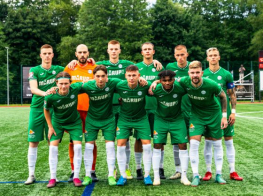 Futbolisti zaļos tērpos sastājušies barā un stāv uz zāliena