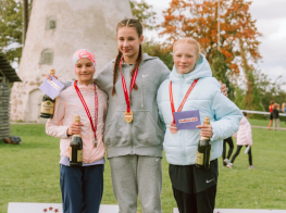 Trīs meitenes stāv uz pjedestāla ar medaļām kaklā