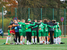 Futbolisti ar komandu aplī stāv un saķērušies futbola laukumā.