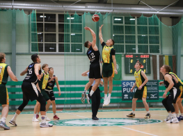 Basketbolisti melnos un dzeltenos kreklos sporta zālē, divi palēkušies gaisā un cīnās par basketbola bumbu