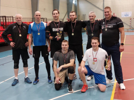 Vairāki vīrieši stāv sporta zālē ar medaļām kaklā