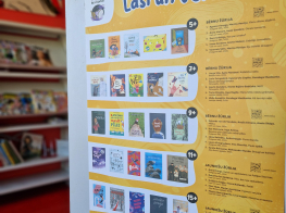 Siena ar dzeltenu plakātu, kur uzzīmētas grāmatas, virsū teksts par grāmatu lasīšanu