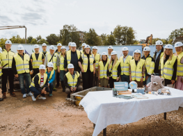 Vairāki cilvēki baltās ķiverēs un dzeltenās vestēs būvlaukumā