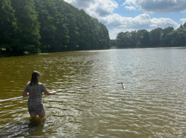 Sieviete peldkostīmā līdz ceļiem iekšā ūdenstilpnē, turot garu kātu ar tvertni galā